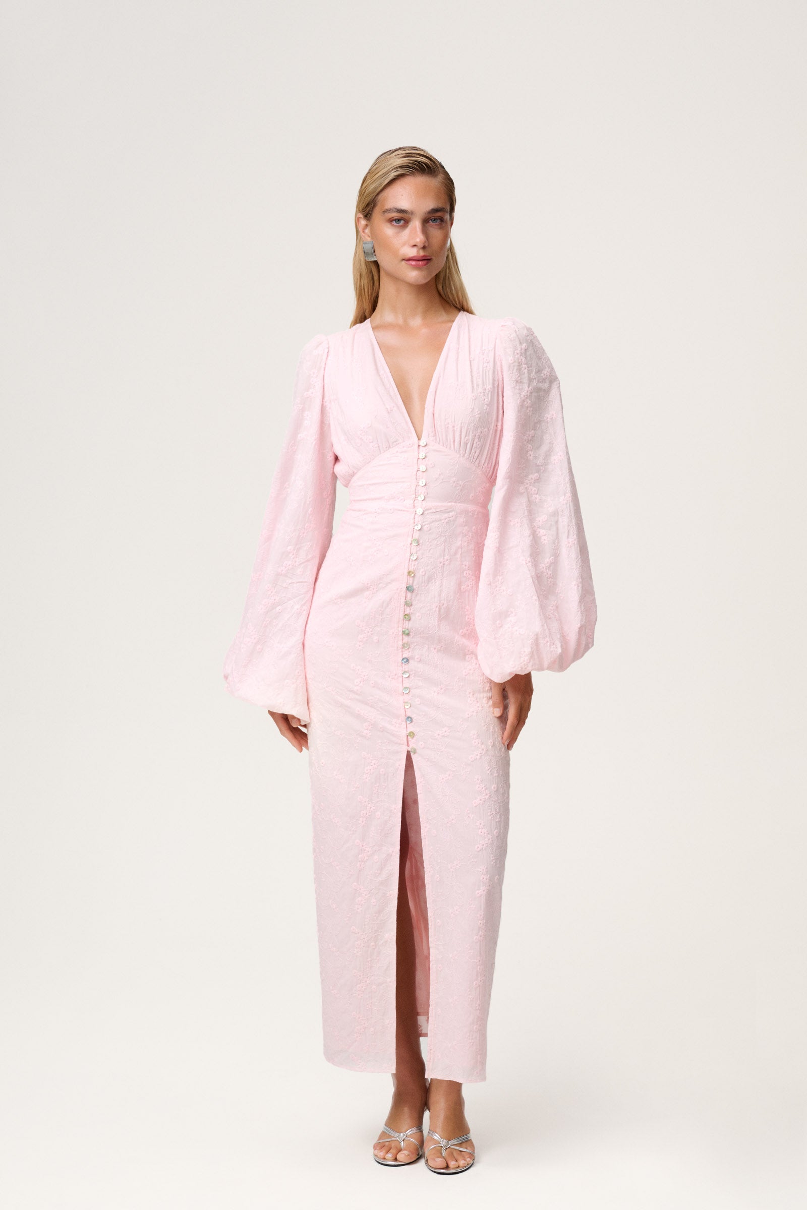 A model wearing a Positano dress , size 36, featured pink long-sleeved maxi dress with embroidery, a V-neck, and front slits. The dress has a hidden zipper at the back and is fully lined. image