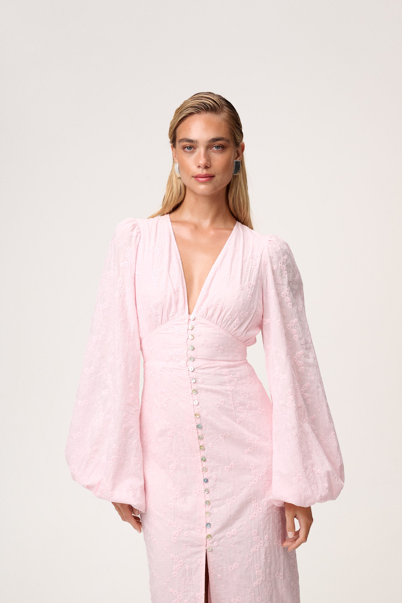 A model wearing a Positano dress , size 36, featured pink long-sleeved maxi dress with embroidery, a V-neck, and front slits. The dress has a hidden zipper at the back and is fully lined. image