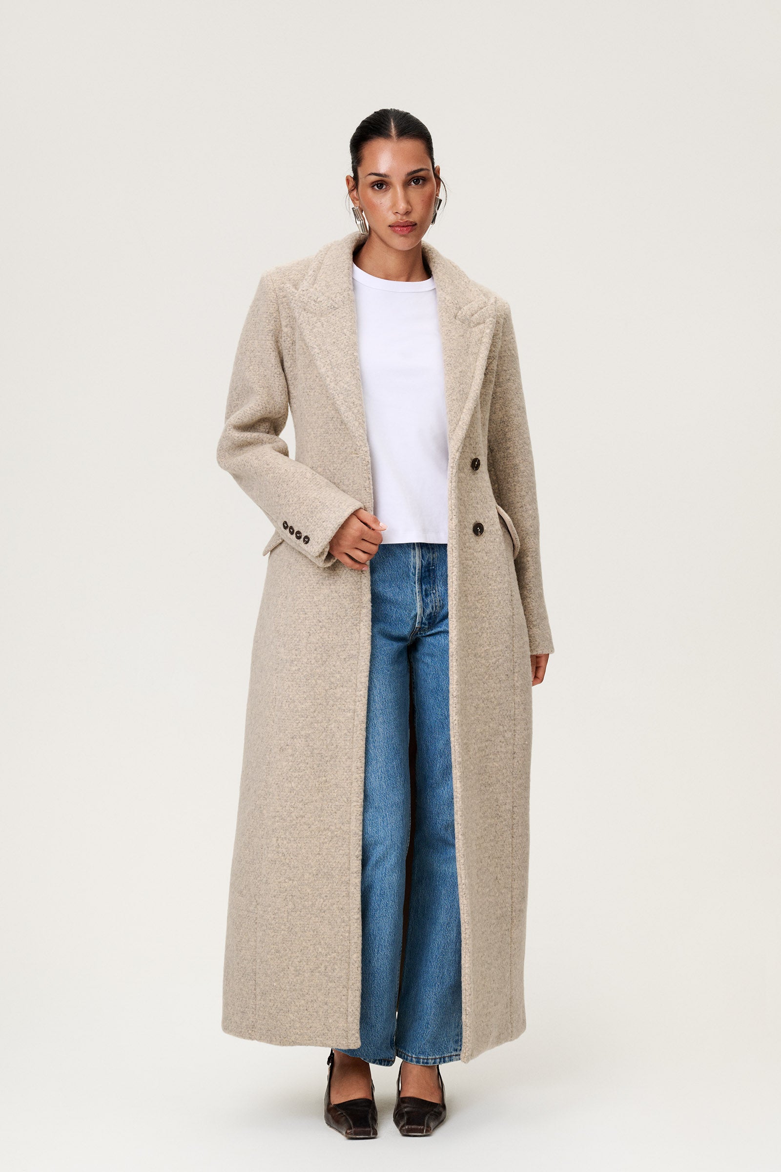 Woman wearing the Stockholm coat in beige melange. A full-length, hourglass coat with a cinched waist, removable suede belt, and structured shoulders. Finished with front pockets and a fully lined interior for warmth and comfort. image