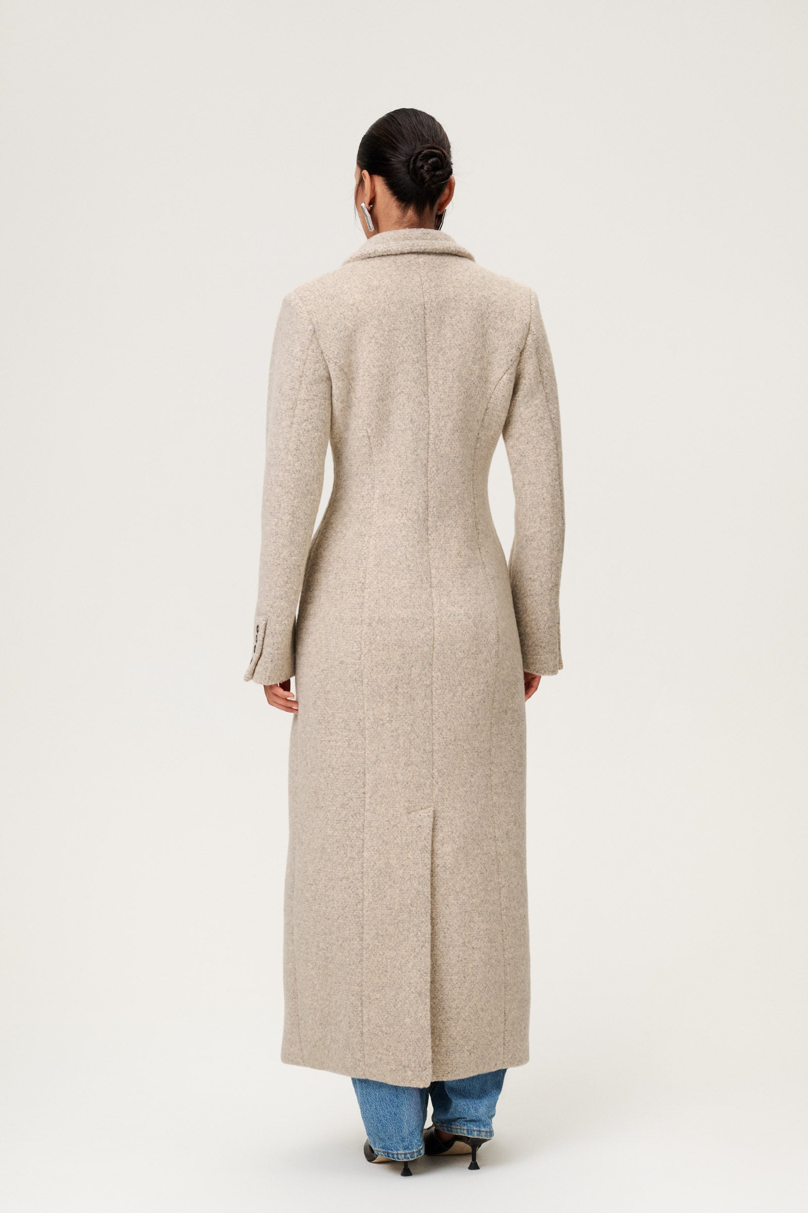 Woman wearing the Stockholm coat in beige melange. A full-length, hourglass coat with a cinched waist, removable suede belt, and structured shoulders. Finished with front pockets and a fully lined interior for warmth and comfort. image
