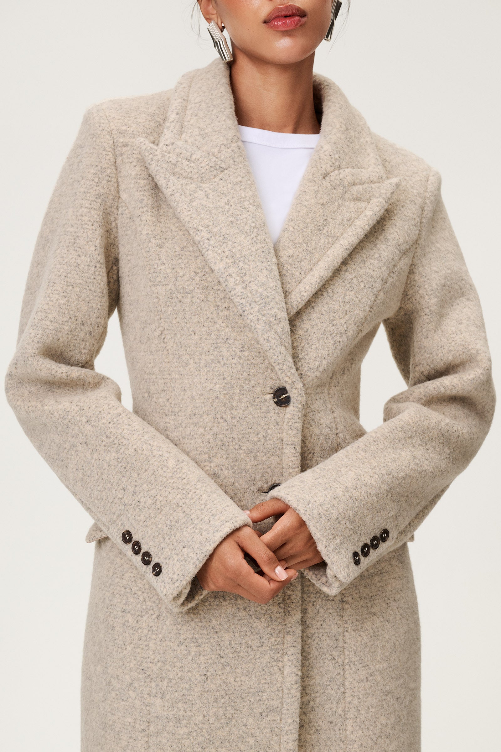 Woman wearing the Stockholm coat in beige melange. A full-length, hourglass coat with a cinched waist, removable suede belt, and structured shoulders. Finished with front pockets and a fully lined interior for warmth and comfort. image