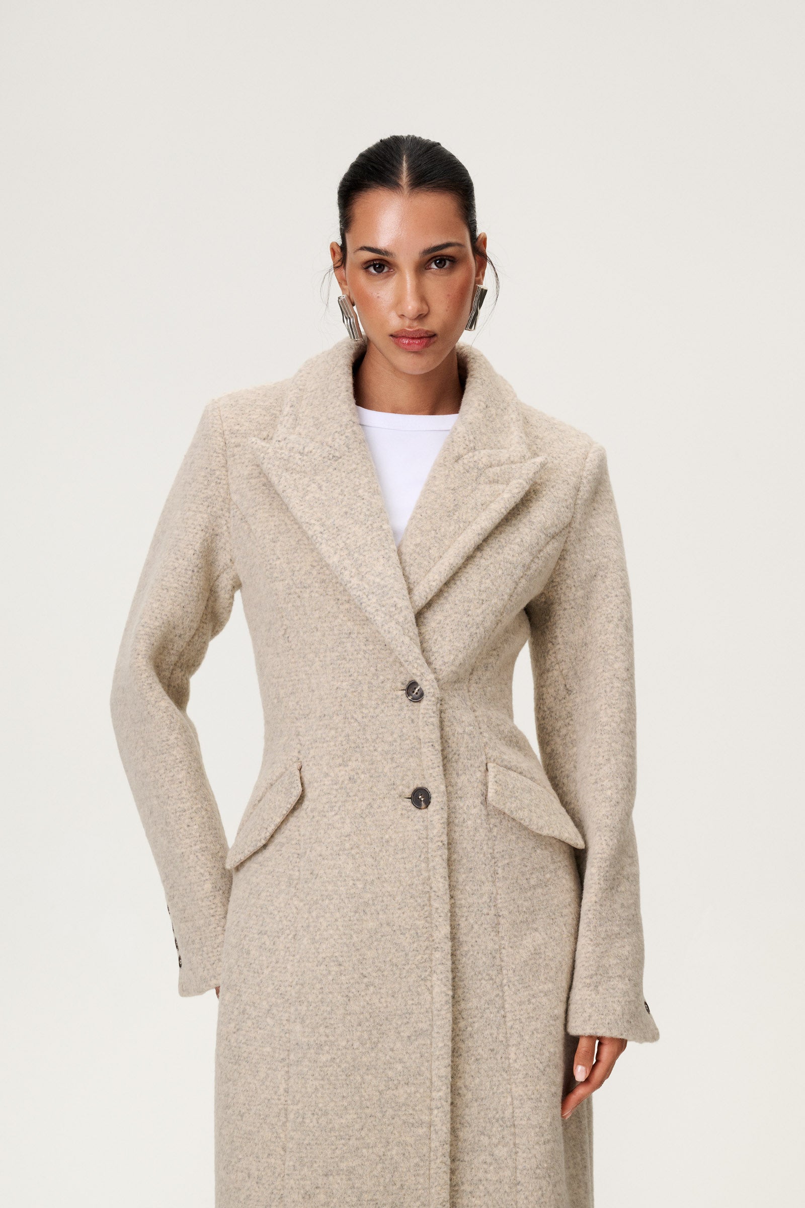 Woman wearing the Stockholm coat in beige melange. A full-length, hourglass coat with a cinched waist, removable suede belt, and structured shoulders. Finished with front pockets and a fully lined interior for warmth and comfort. image