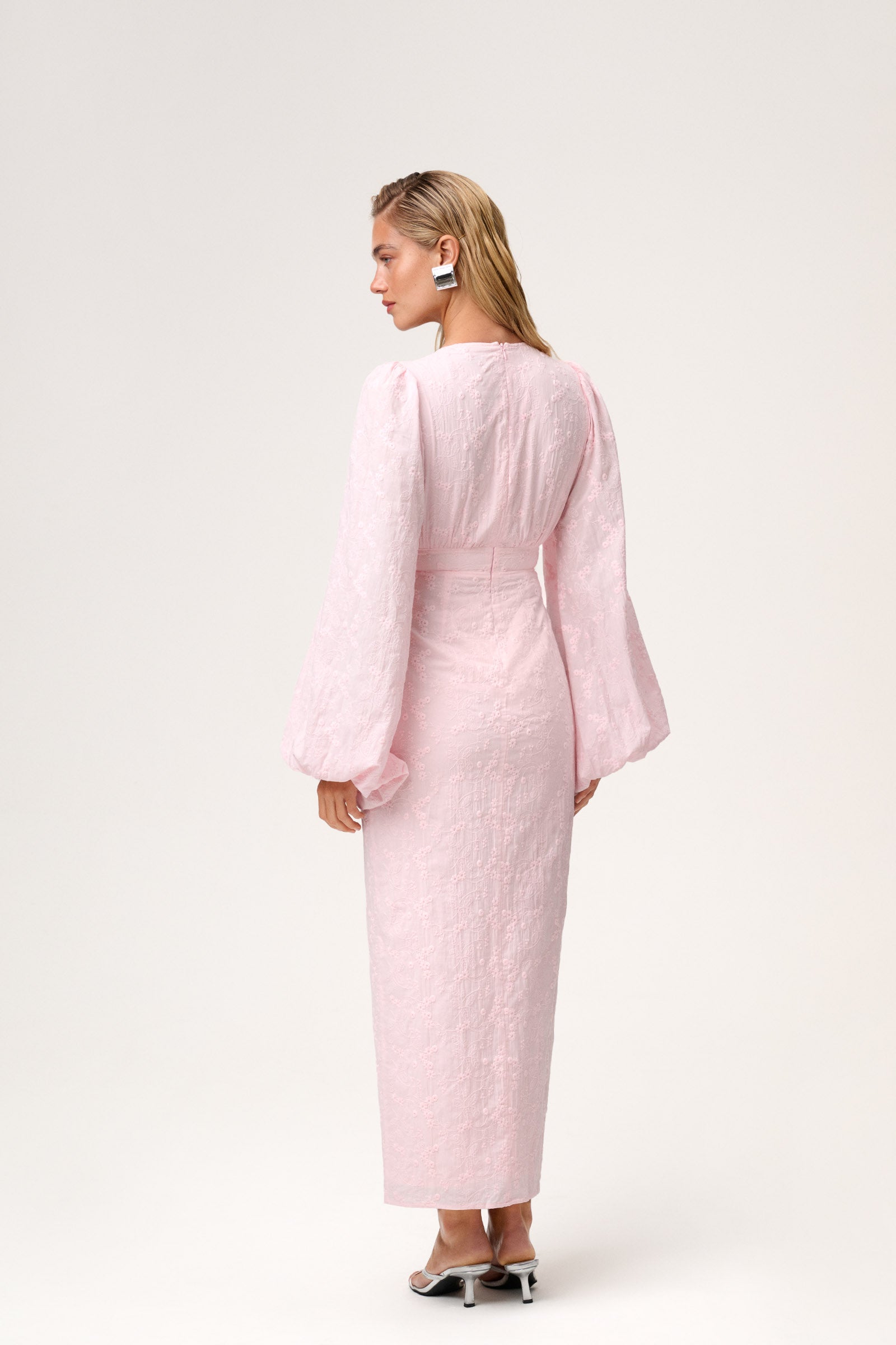 A model wearing a Positano dress , size 36, featured pink long-sleeved maxi dress with embroidery, a V-neck, and front slits. The dress has a hidden zipper at the back and is fully lined. image