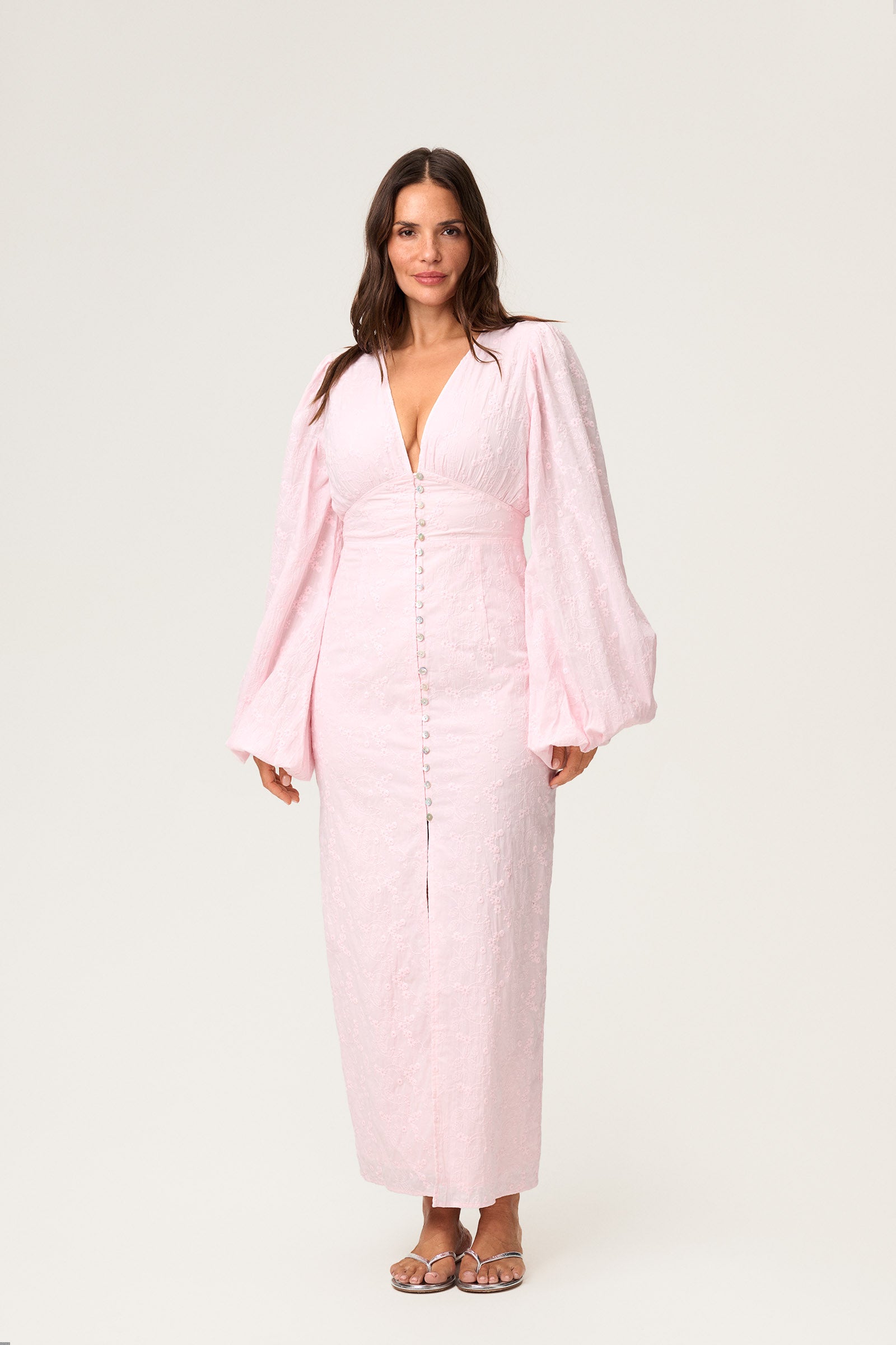 A model wearing a Positano dress , size 42, featured pink long-sleeved maxi dress with embroidery, a V-neck, and front slits. The dress has a hidden zipper at the back and is fully lined. image