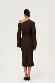 Woman wearing the Arona knitted midi dress in brown melange with one padded shoulder, full-length sleeves, a back slit, and a heavy cable-knit design. thumbnail image