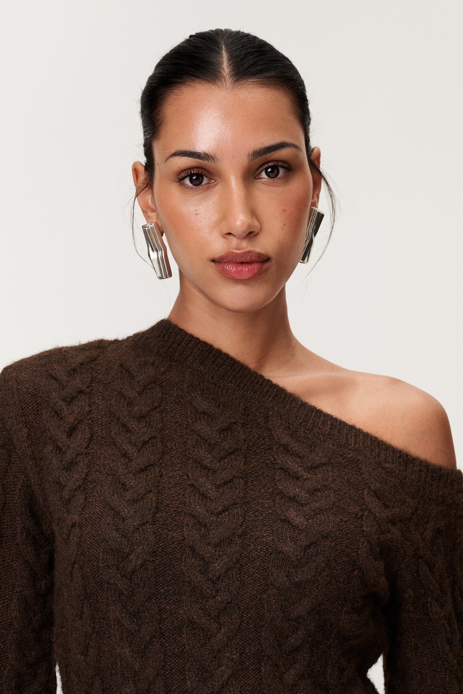 Woman wearing the Arona knitted midi dress in brown melange with one padded shoulder, full-length sleeves, a back slit, and a heavy cable-knit design.Woman wearing the Arona knitted midi dress in brown melange with one padded shoulder, full-length sleeves, a back slit, and a heavy cable-knit design. image