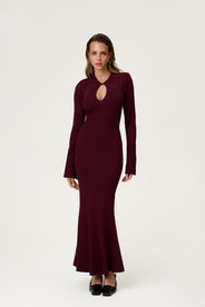 Woman wearing the Basel dress in burgundy, a knitted maxi with a collared cut-out neckline, decorative seams, and a flared hem. Finished with long sleeves and wide cuffs. thumbnail image