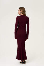 Woman wearing the Basel dress in burgundy, a knitted maxi with a collared cut-out neckline, decorative seams, and a flared hem. Finished with long sleeves and wide cuffs. thumbnail image