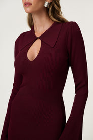 Woman wearing the Basel dress in burgundy, a knitted maxi with a collared cut-out neckline, decorative seams, and a flared hem. Finished with long sleeves and wide cuffs. thumbnail image