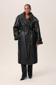 A person standing wearing a black Manchester coat with oversized fit, midi length, faux shearling lining, and a belt at the waist with D-buckle. thumbnail image