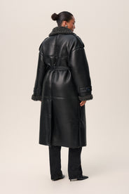A person standing wearing a black Manchester coat with oversized fit, midi length, faux shearling lining, and a belt at the waist with D-buckle. thumbnail image