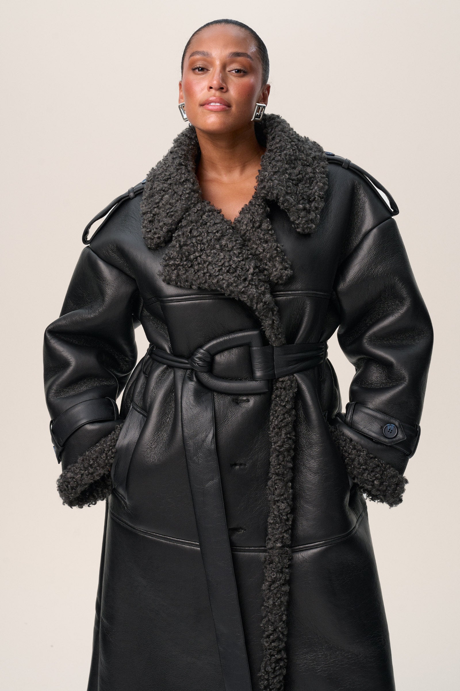 A person standing wearing a black Manchester coat with oversized fit, midi length, faux shearling lining, and a belt at the waist with D-buckle. image