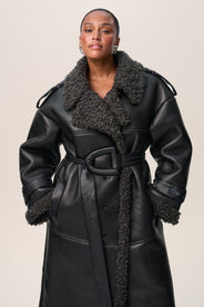 A person standing wearing a black Manchester coat with oversized fit, midi length, faux shearling lining, and a belt at the waist with D-buckle. thumbnail image