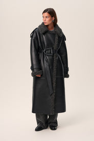 A person standing wearing a black Manchester coat with oversized fit, midi length, faux shearling lining, and a belt at the waist with D-buckle. thumbnail image