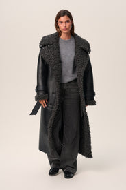 A person standing wearing a black Manchester coat with oversized fit, midi length, faux shearling lining, and a belt at the waist with D-buckle. thumbnail image