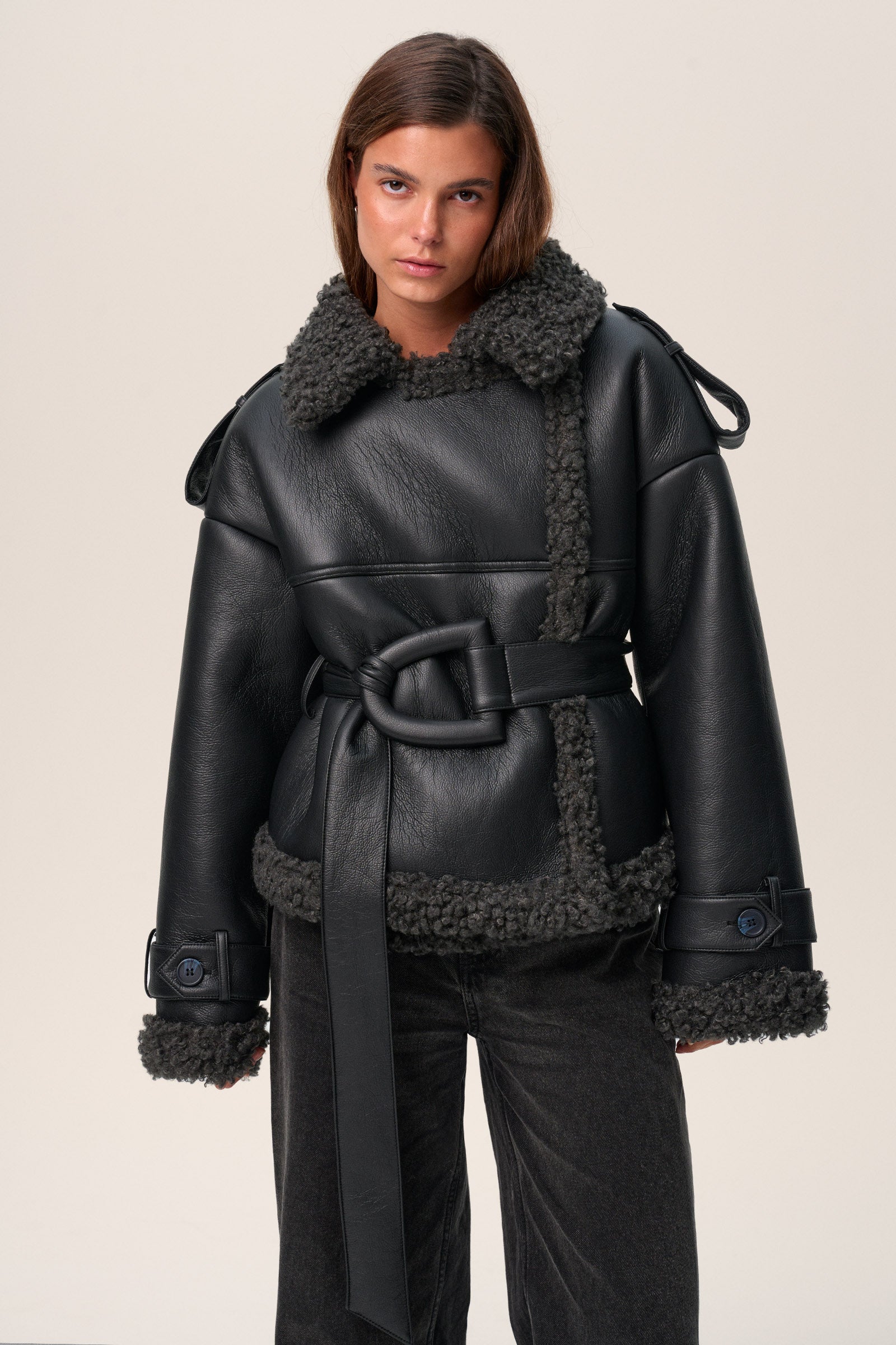A model wearing a black Manchester cropped jacket with oversized fit, faux shearling lining, side pockets, and a belt with a D-shaped buckle. image