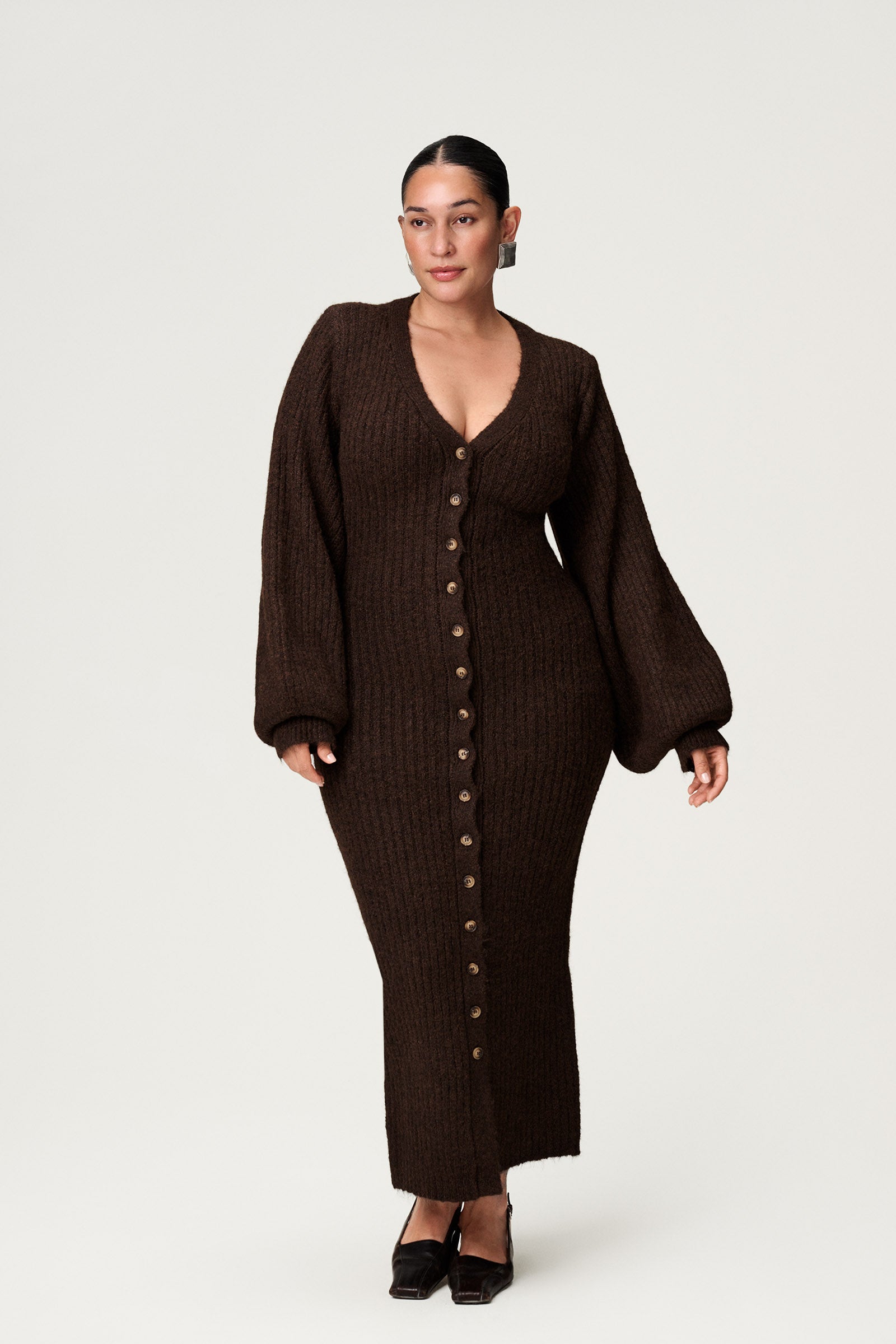 Woman wearing a brown knitted dress with button details, and ballon sleves on a white background. Styled with black low heels.  image