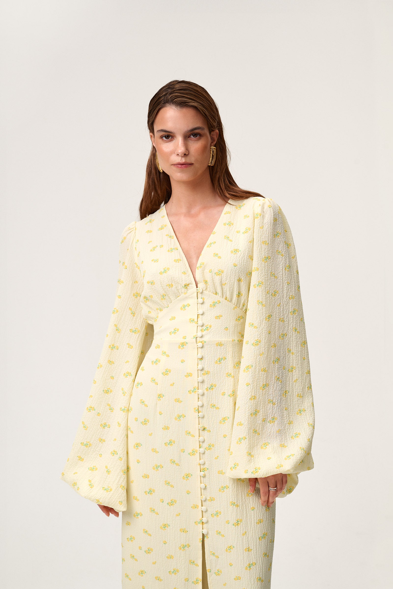 A woman wearing Positano dress, a pastel yellow maxi dress with long sleeves, a V-neck collar, buttons and slits at the front, and a hidden zipper at the back.
 image