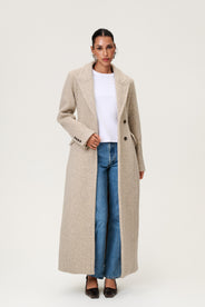 Woman wearing the Stockholm coat in beige melange. A full-length, hourglass coat with a cinched waist, removable suede belt, and structured shoulders. Finished with front pockets and a fully lined interior for warmth and comfort. thumbnail image