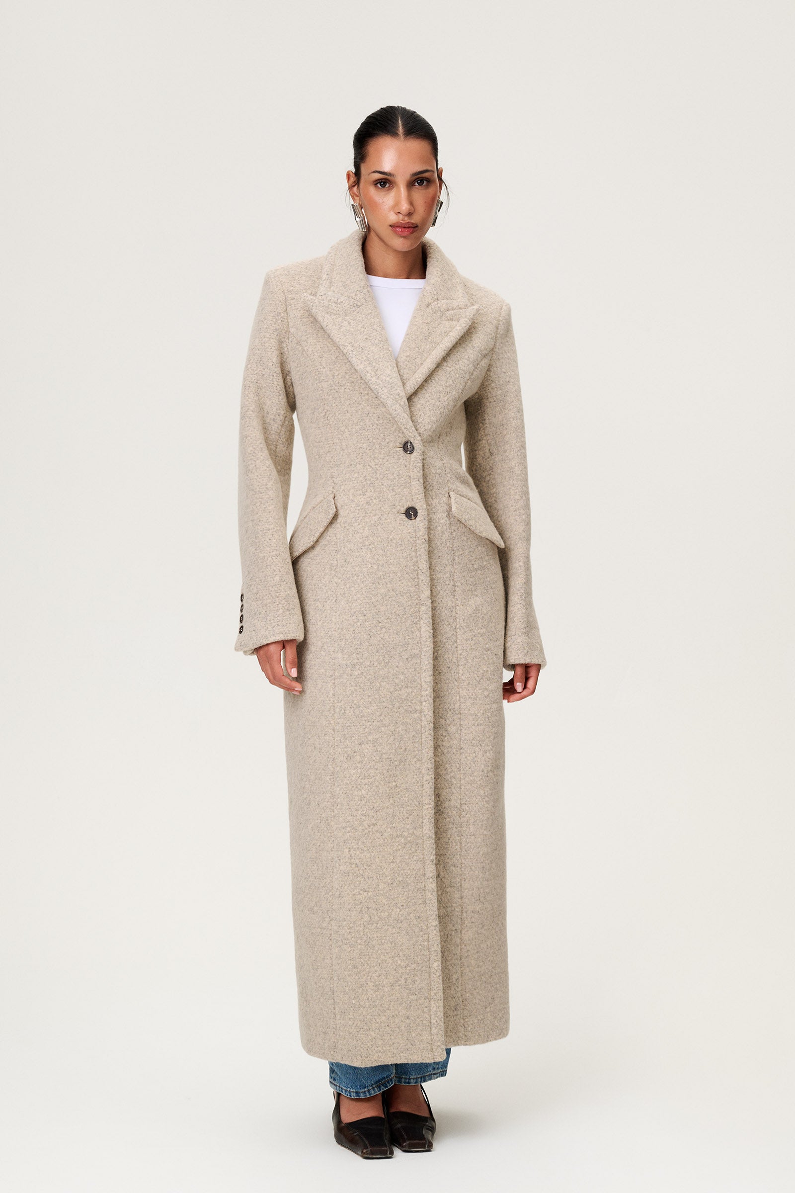 Woman wearing the Stockholm coat in beige melange. A full-length, hourglass coat with a cinched waist, removable suede belt, and structured shoulders. Finished with front pockets and a fully lined interior for warmth and comfort. image