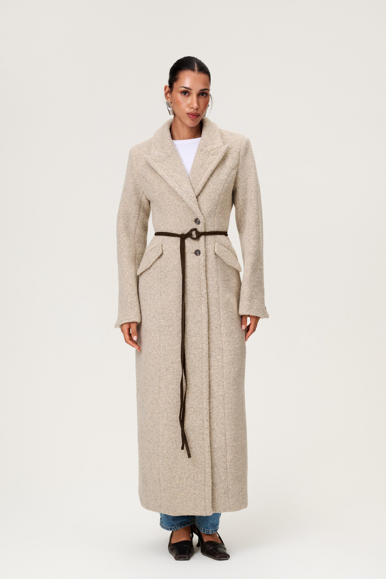 Woman wearing the Stockholm coat in beige melange. A full-length, hourglass coat with a cinched waist, removable suede belt, and structured shoulders. Finished with front pockets and a fully lined interior for warmth and comfort. image